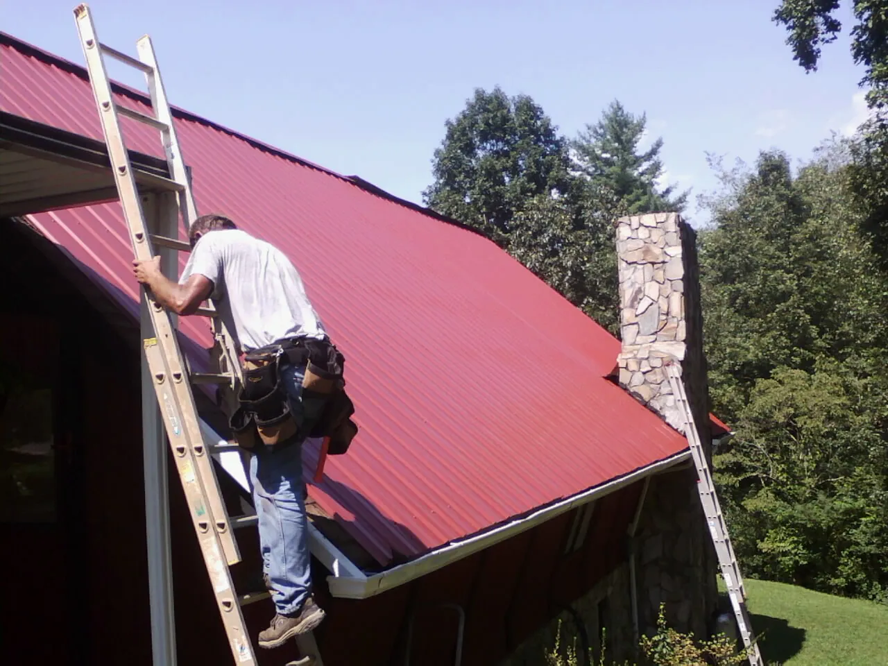 Roofer on a ladder beside a red metal roof and stone chimney — Soffit & Fascia Repair by Roofing Brothers in Glenside
